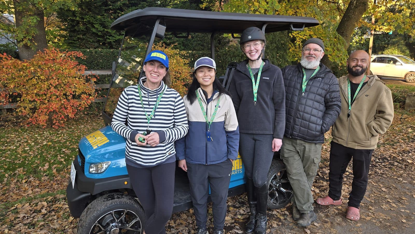 Five people wearing lanyards standing beside a HeyYa cart among autumn leaves
